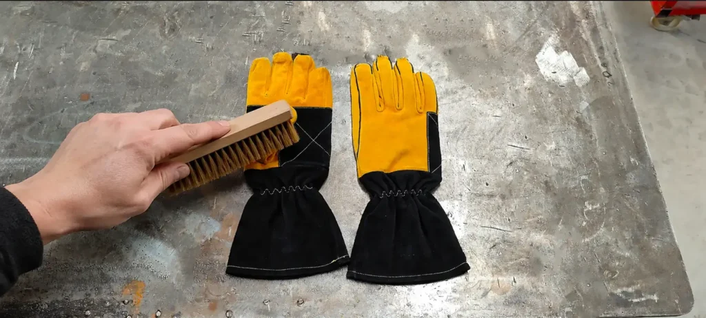 Cleaning ARASWELD leather welding gloves with a soft brush to remove dust, slag, and welding debris before conditioning and maintenance.