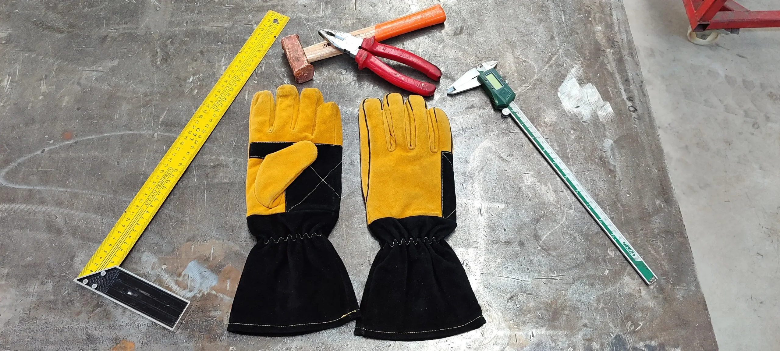 arasweld Professional welding glove with Premium Leather reinforced palm and split leather protection, Sewn with Kevlar thread Arasweld. Which is located on a welding workbench, with a hand-held pliers, a ruler. and a welded piece next to it. By a professional welder who used Arasweld gloves.